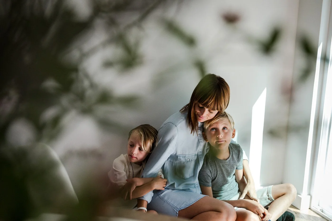 Mom cuddles with son and daughter as they lean against a white wall, shot at home through a bouquet flowers.