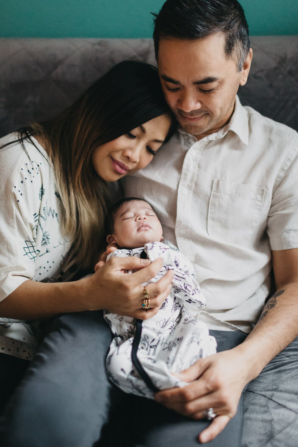 Dad hold newborn daughter in lap while mom leans into him and gazes at baby.