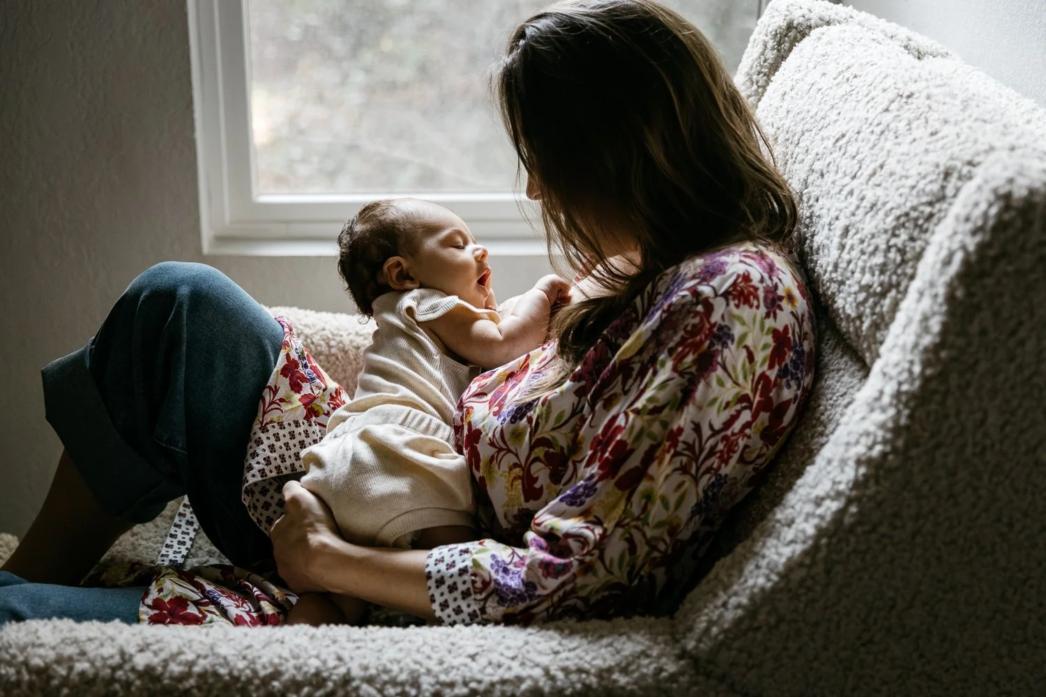 Mom in colorful robe, sits in cozy chair in front of window and gazes at newborn baby, shot during newborn session at home in Oakland.