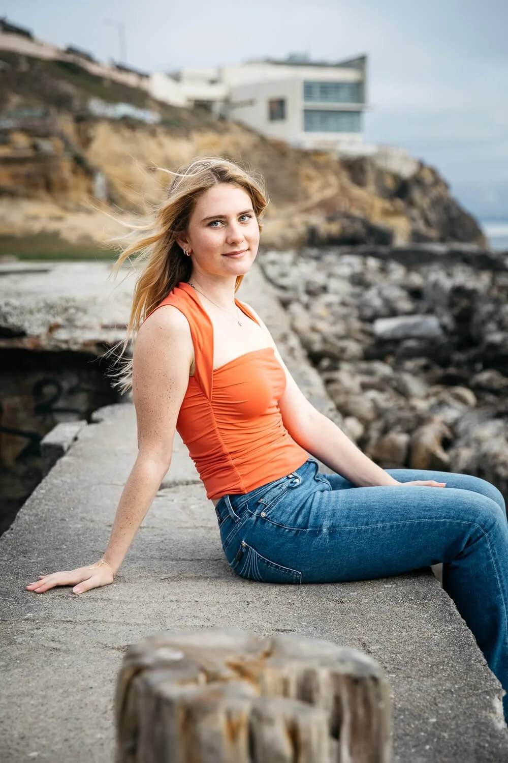 Senior girl in salmon-colored tank and jeans sits on stone wall at Sutro Baths in San Francisco and smiles at the camera.