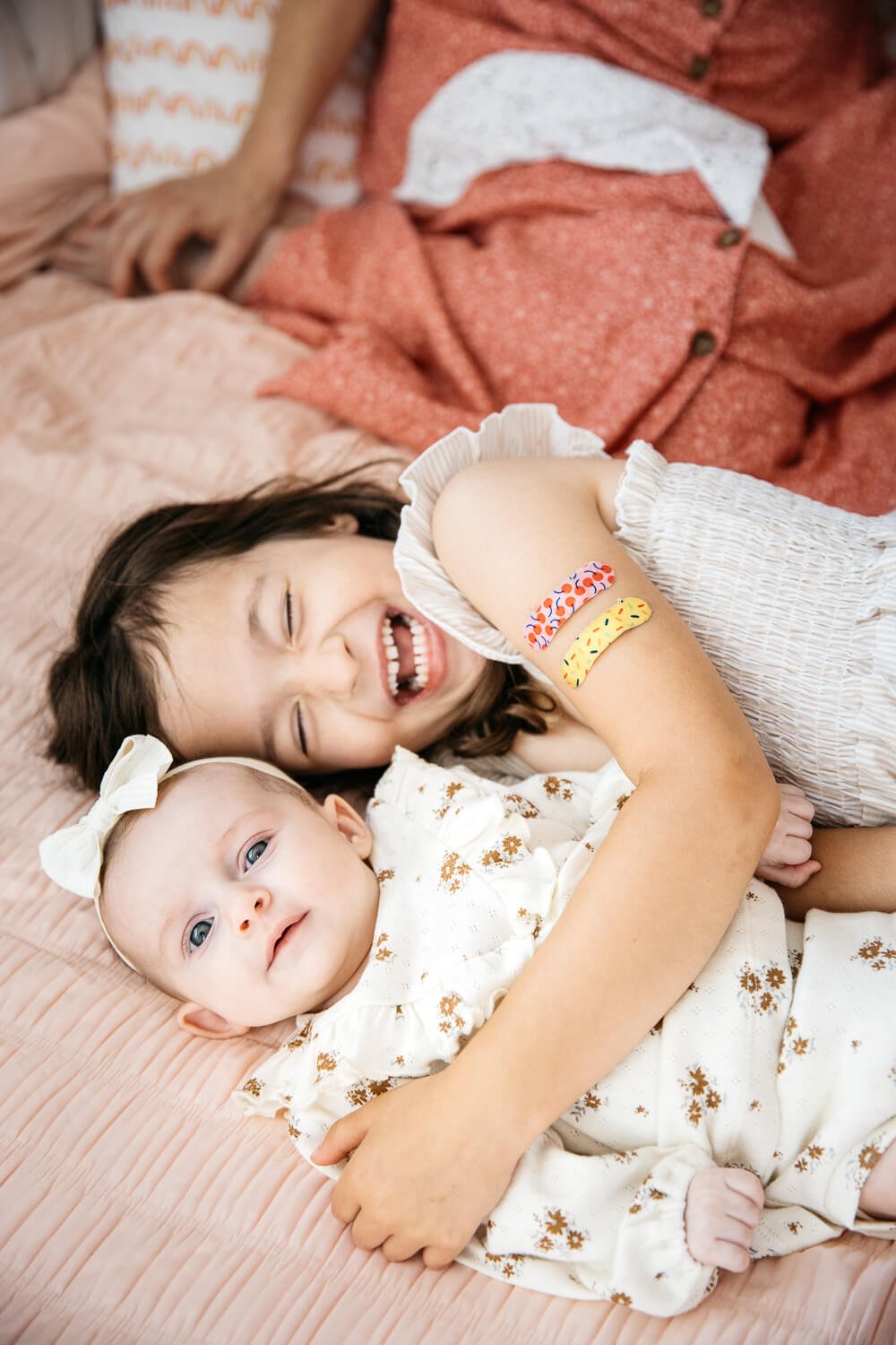 Big sister holds newborn baby on pink bedspread and laughs.