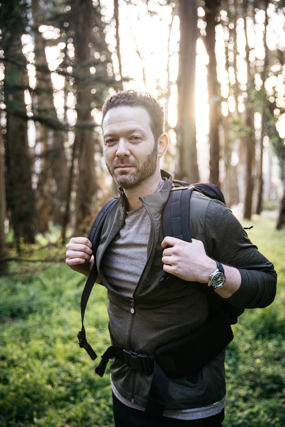 Man in half-zip and backpack stands in eucalyptus grive with the sun setting behind him.