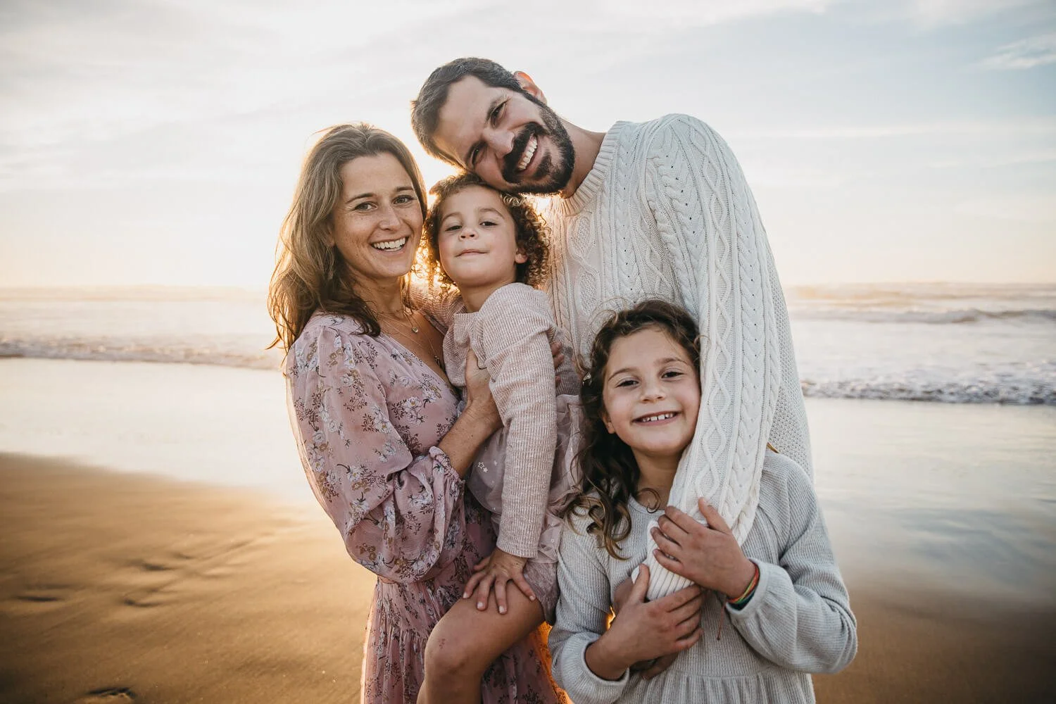 Family of four stand at water line at Ocean Beach in San Francisco, as sun sets behind them and creates peachy glow.