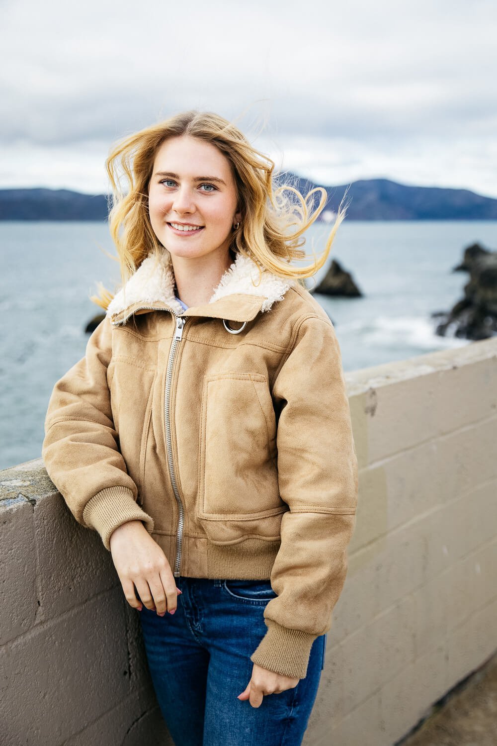 Teen girl wearing beige shearling bomber jacket and jeans, stands at cliff's edge at Lands End while wind blows her hair.