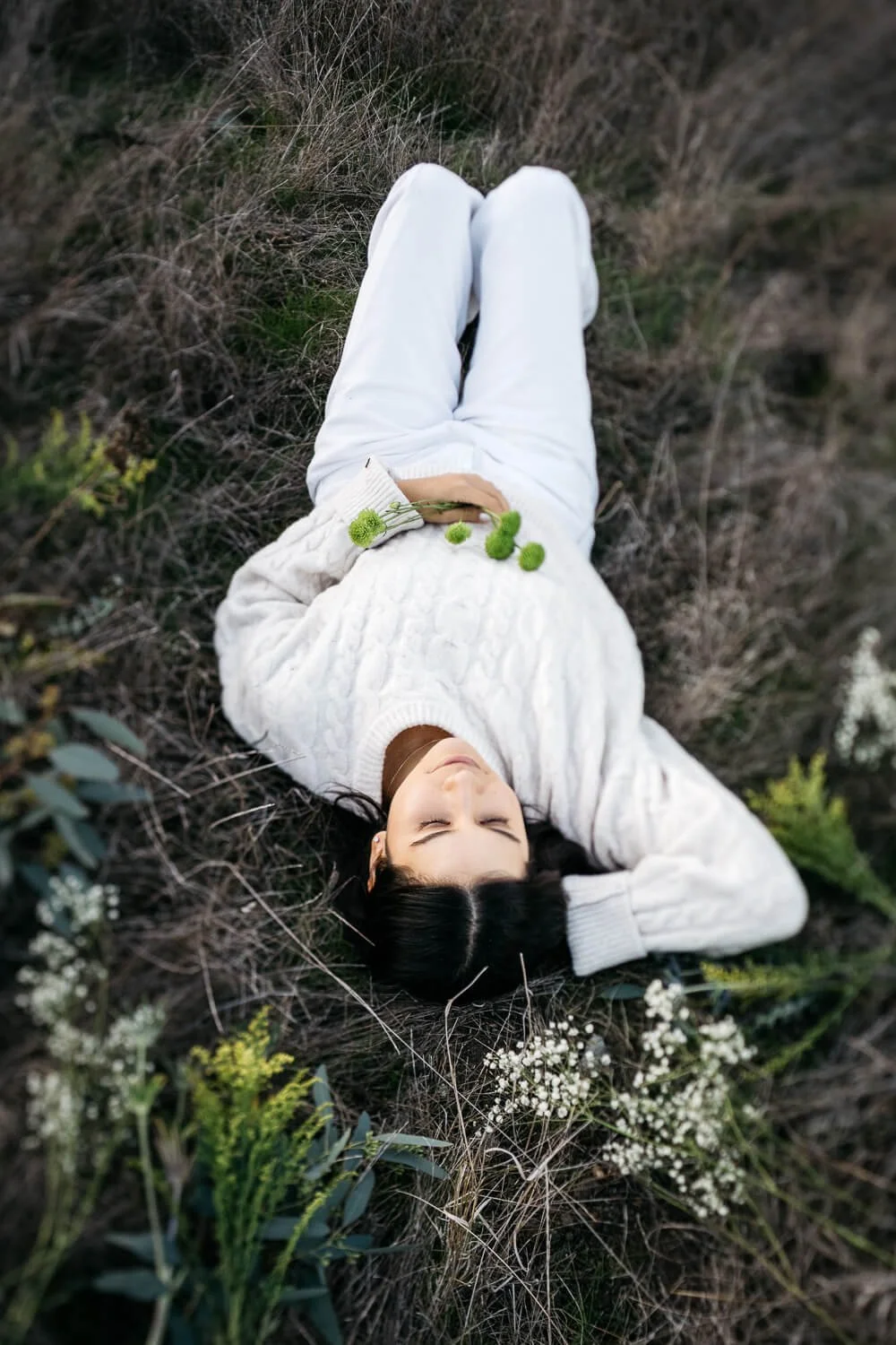 Woman in white sweater and pants lies in field of flowers at sunset.