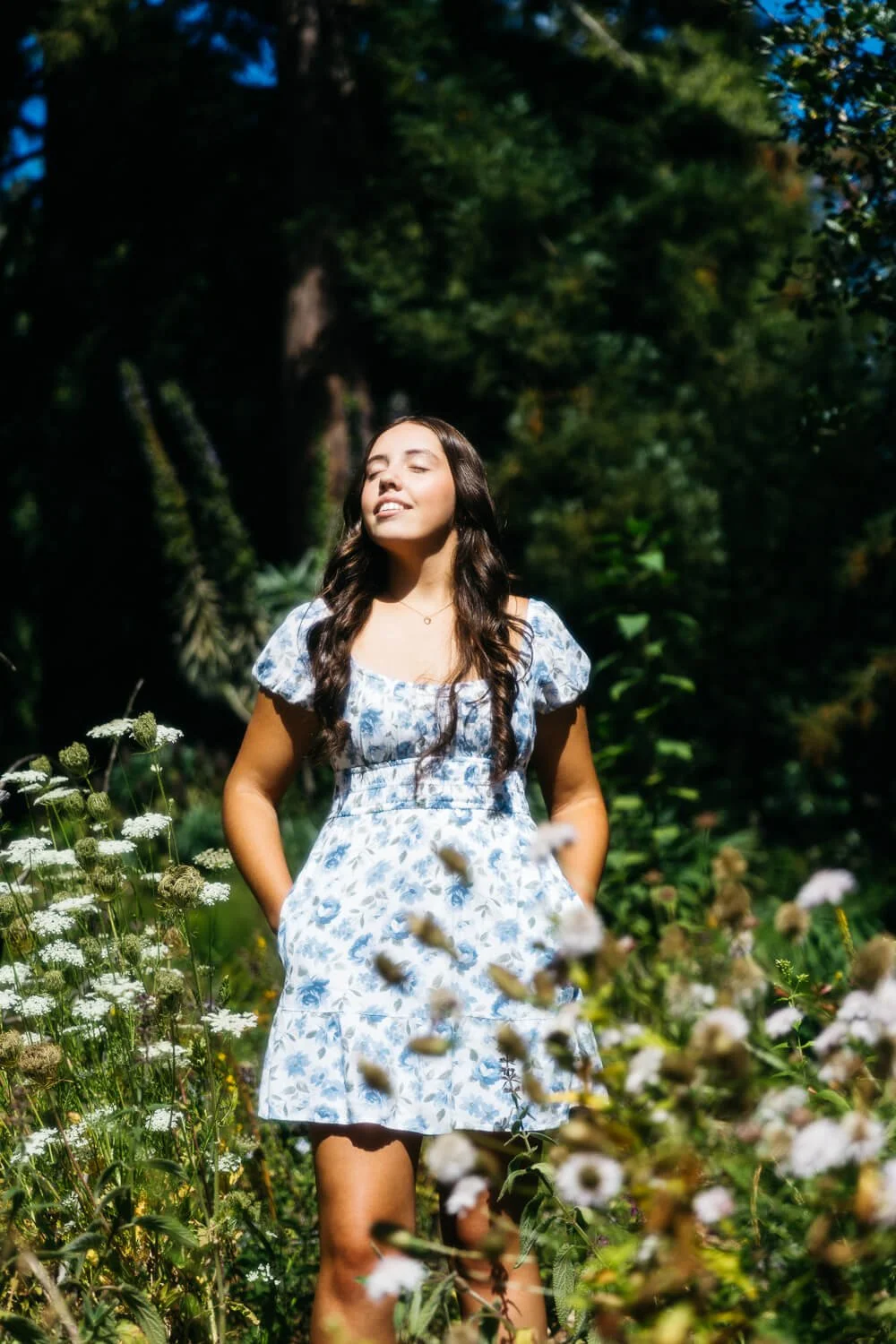 Teen with long brown hair and white dress with blue flowers stands among wildflowers during senior session at Blake Garden.