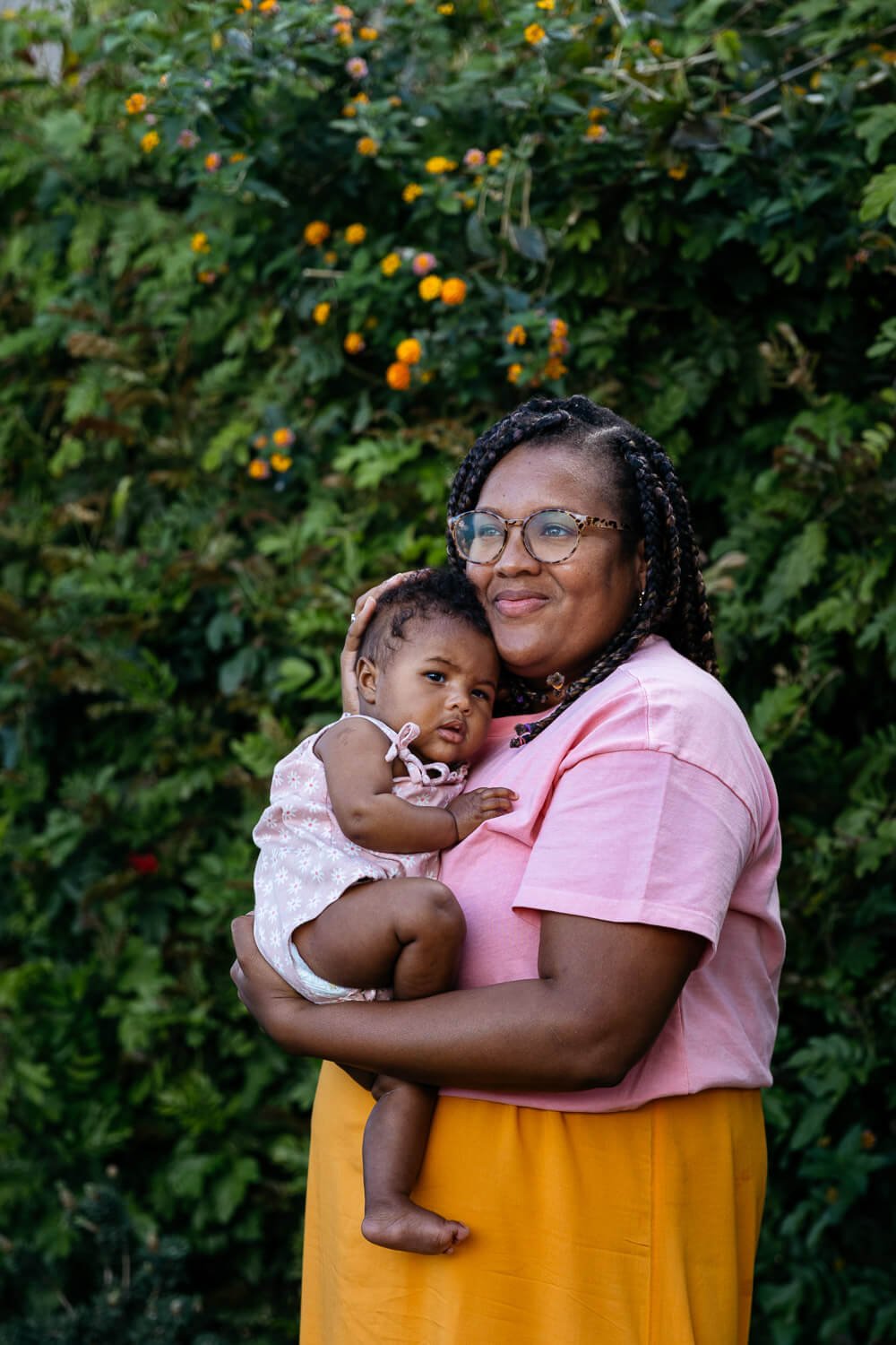 Smiling mom wearing pink and orange dress stands in front of orange tree cradling baby daughter.
