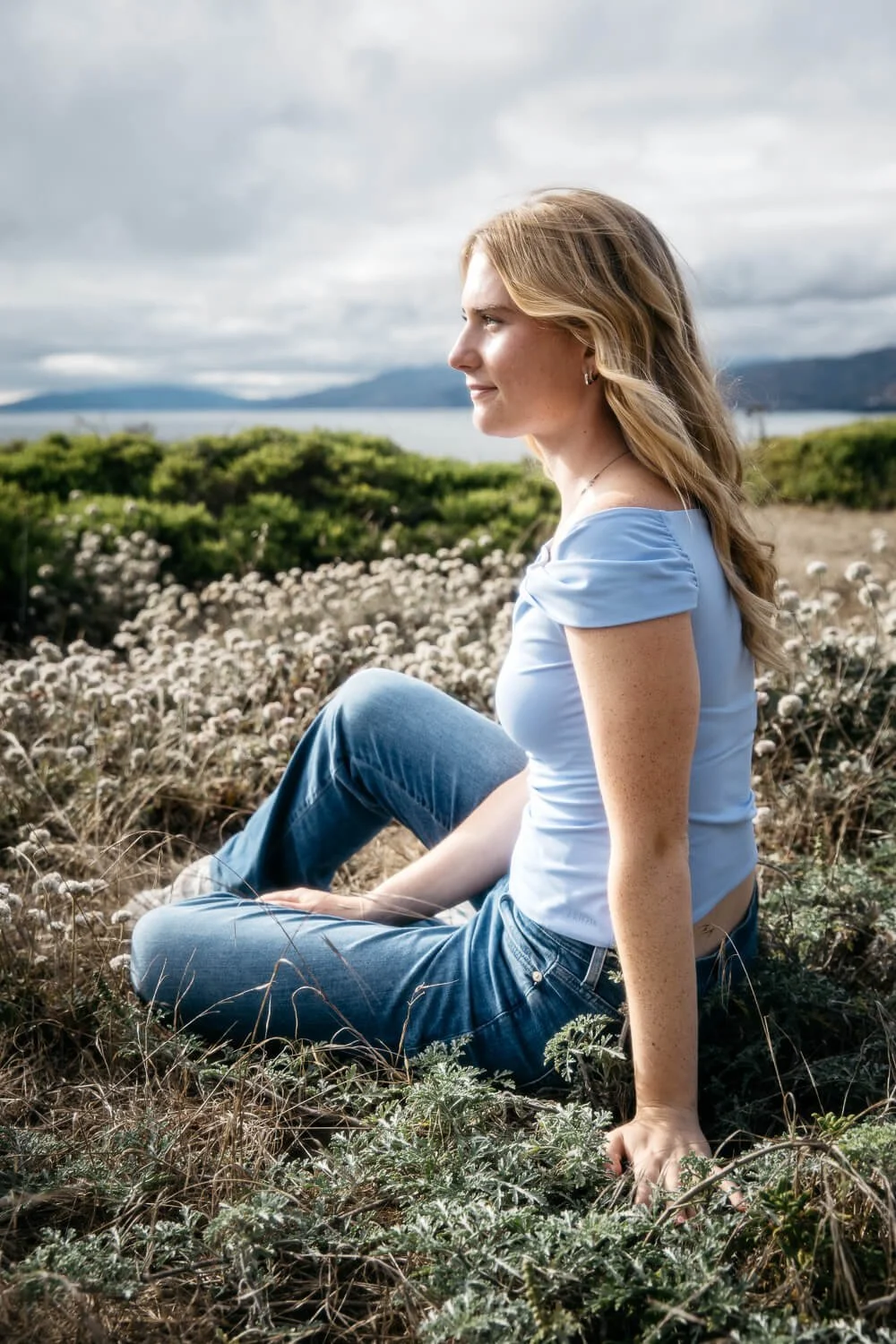 Teen girl wearing blue tanks top and jeans sits in coastal scrub plants at Lands End and looks out to sea.