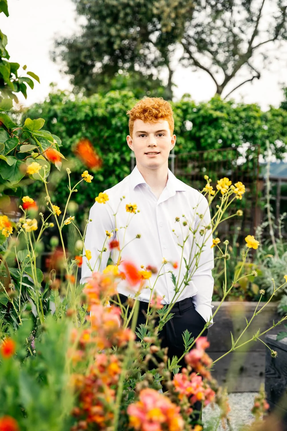 Senior portrait of redheaded boy wearing white button up shirt, standing amongst orange and yellow flowers in his backyard, shot in Orinda, CA.
