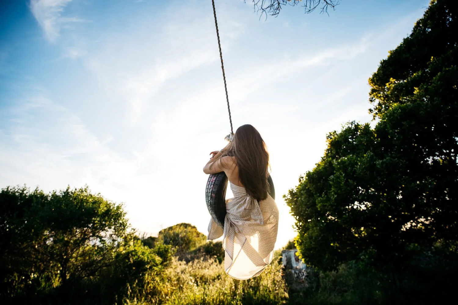 Senior girl in long gingham dress swings on tire swing with sunlight backlighting her and shining through her skirt, shot during senior photo session at Albany Bulb.