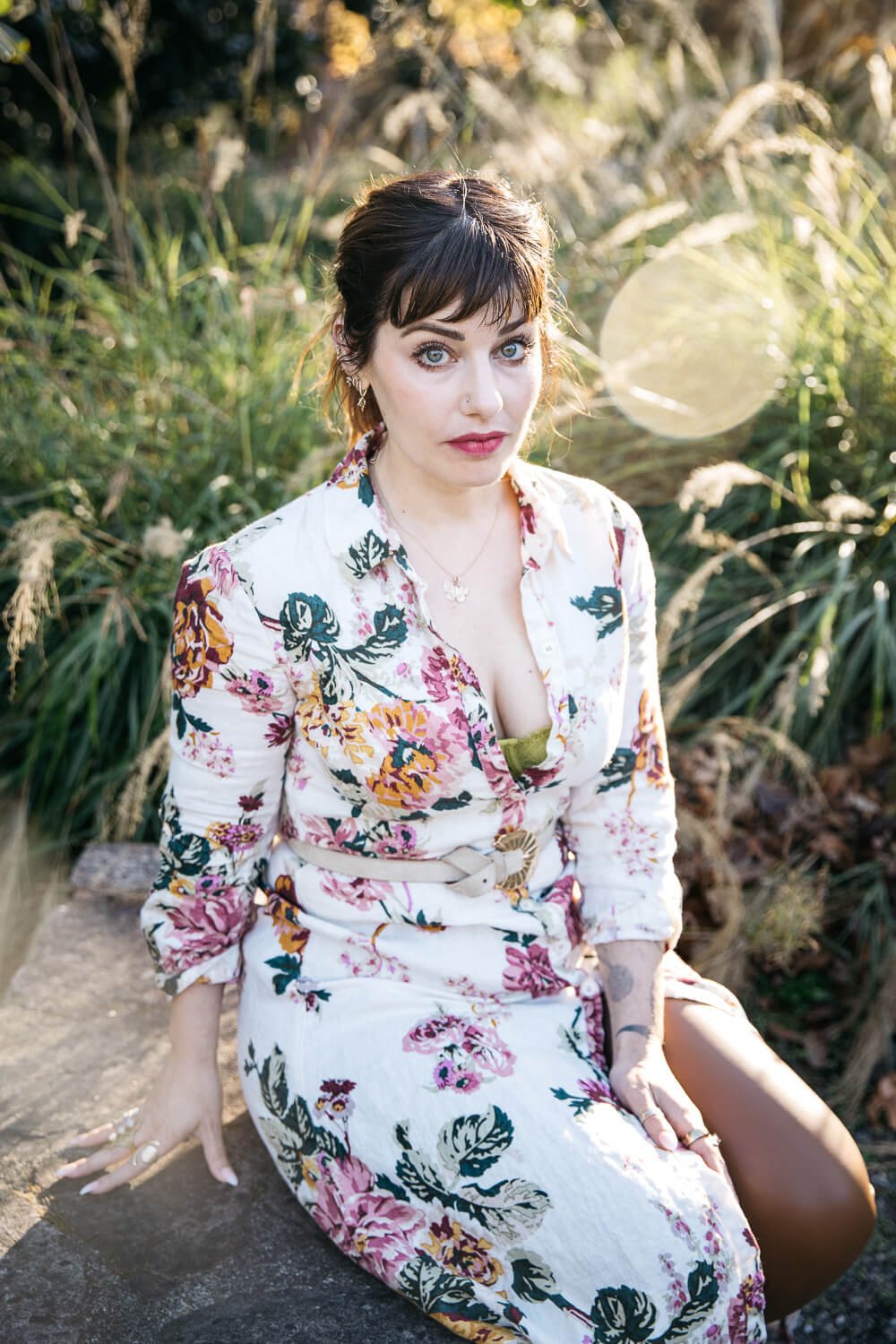 Woman in white floral dress sits among tall grasses, backlit with sun flare.