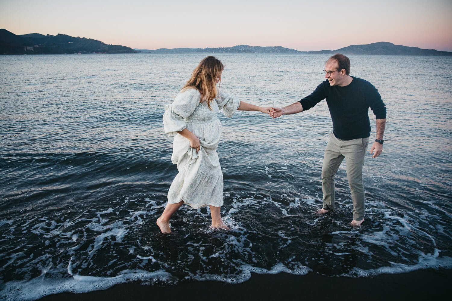 Man and women dance in the surf during maternity session at Crissy Field Beach at sunset.