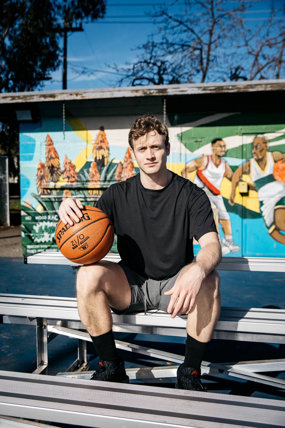Man in black t-shirt, sits on bleachers in front of community mural, holding a basketball.
