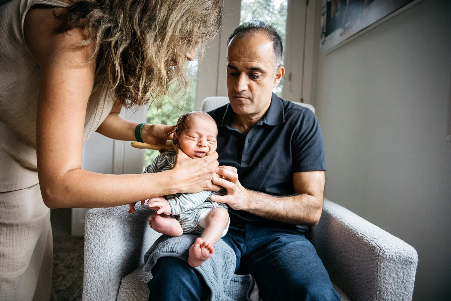 Dad holds newborn baby in his lap, a burp cloth on his knee. Mom bends over and cradles the infants face while she burps him. Moment captured during in home newborn lifestyle session.