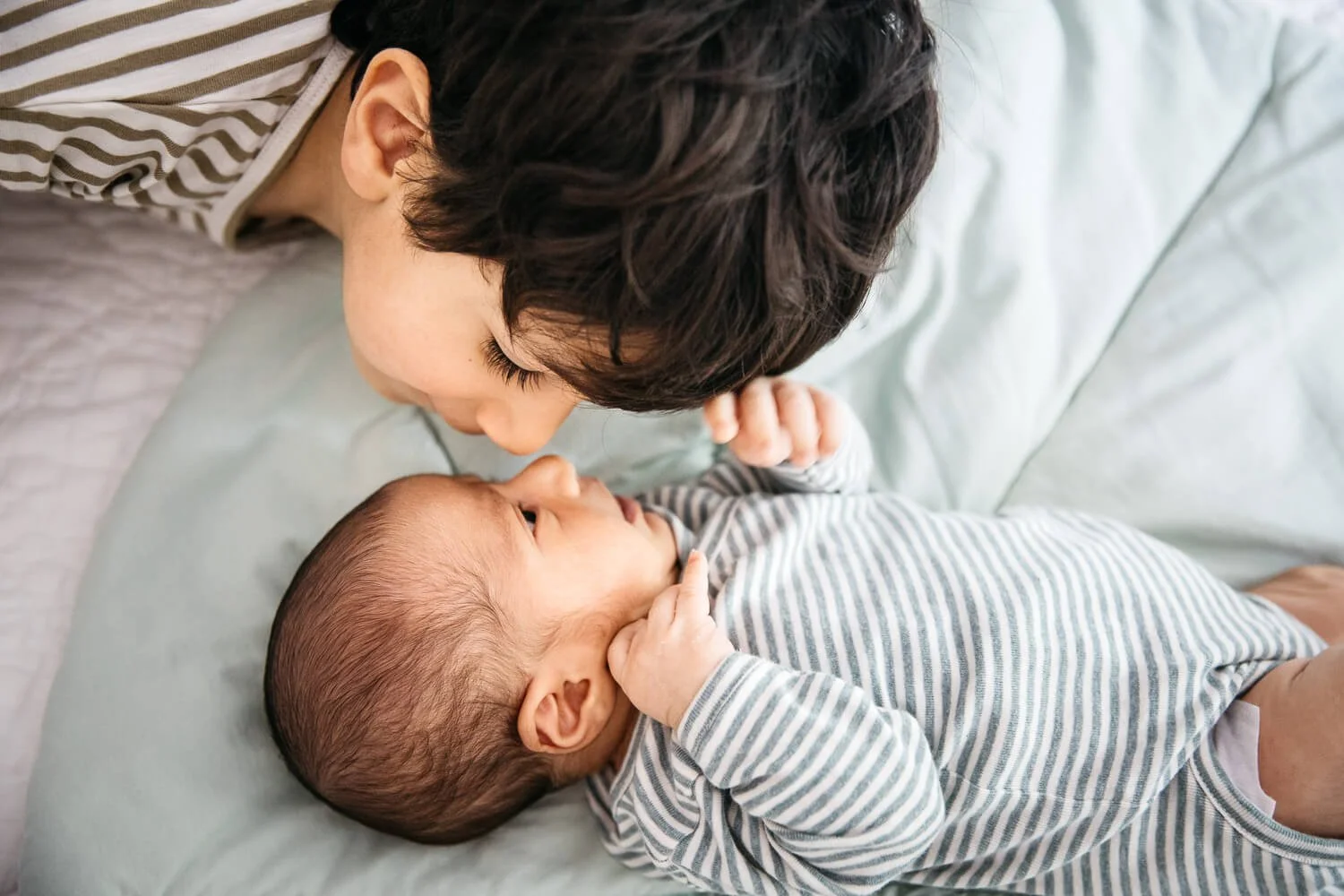 Close portrait of toddler big brother face to face with newborn baby brother, noses almost touching as they look into each other's eyes. Taken during in home newborn lifestyle photo shoot.