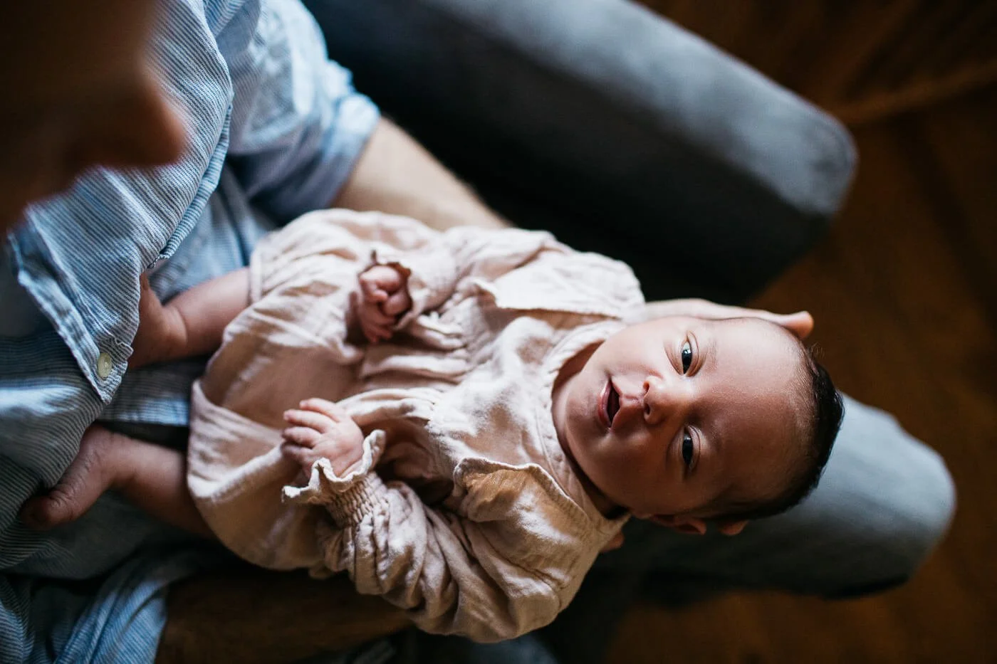 Overhead shot of newborn baby girl in pink dress, smiling at camera while sitting in dad's lap.