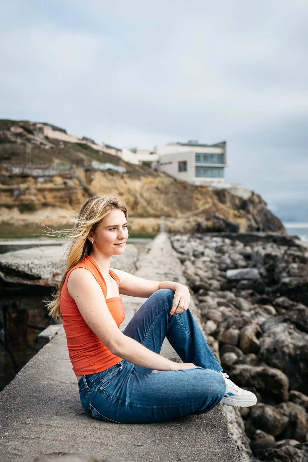 Senior girl in salmon-colored tank and jeans sits on stone wall at Sutro Baths in San Francisco and looks out at the water.
