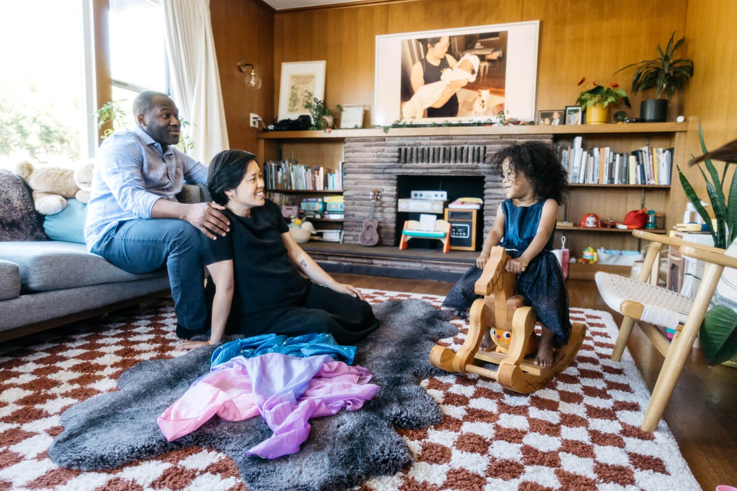 Pregnant Mom and dad sit together in living room and chat with toddler daughter who sits on a rocking horse, shot during a family lifestyle photo session at home in Berkeley.