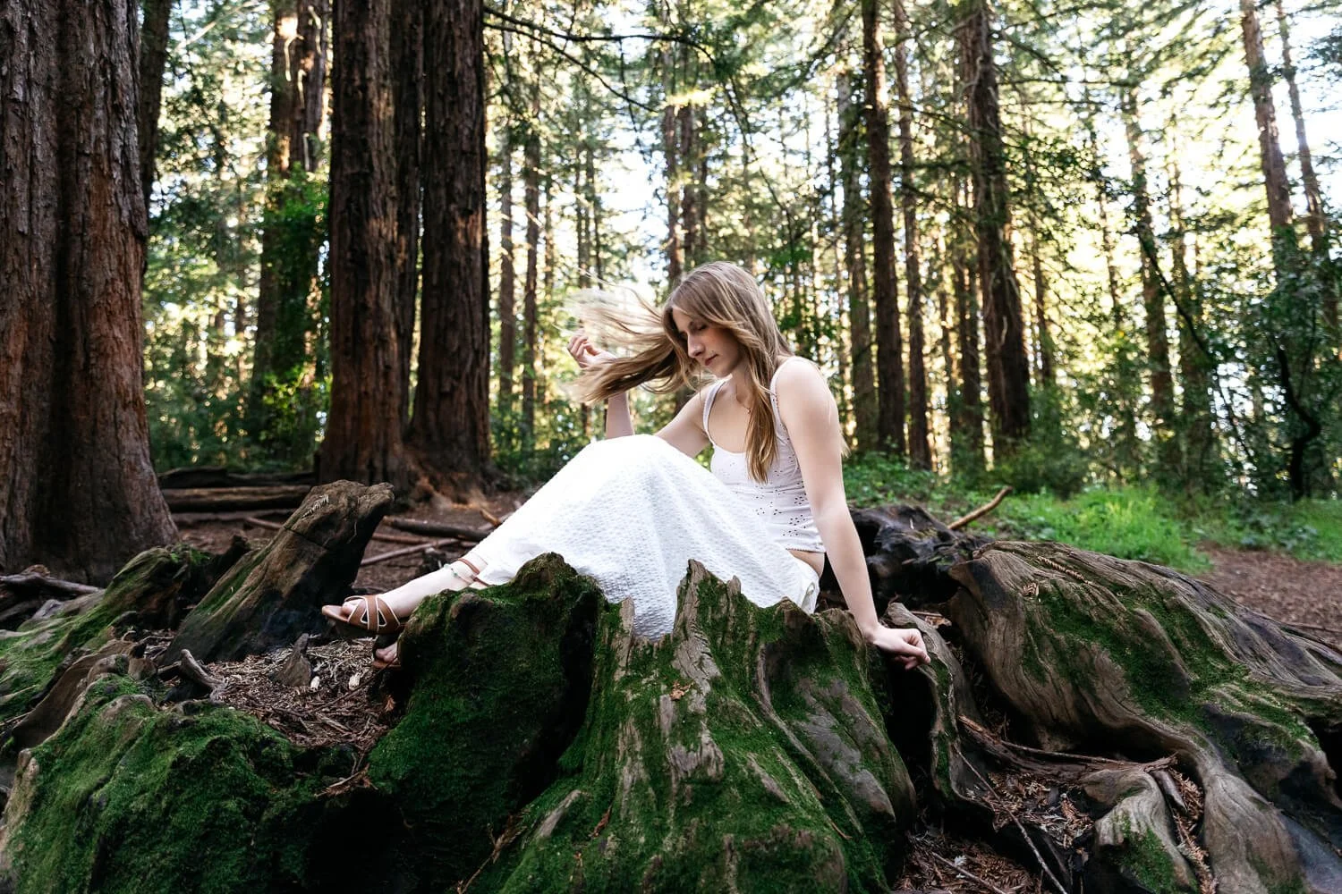 Teen in white skirt and tank top sits in redwood fairy ring and flips her hair. Shot during senior photo session at Redwood Regional in Oakland.