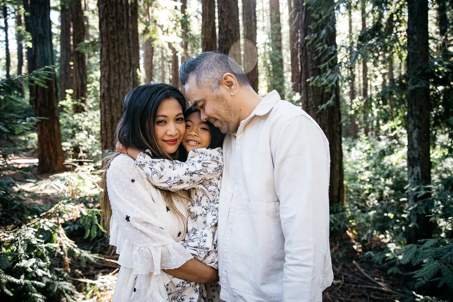 Family of 3 wearing black and white pose in family hug in a redwood grove. The mother and daughter smile at the camera and the father is turned into them. The pose makes them look naturally connected.