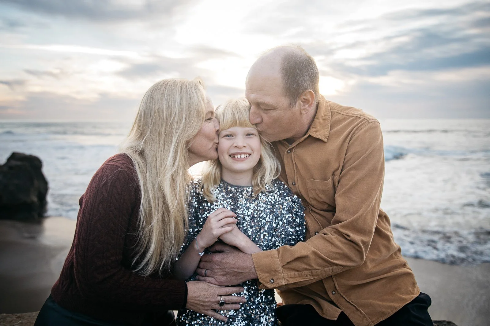 Mom and Dad kiss tween daughter in silver sequin dress while sitting at Sutro Baths in San Francisco at sunset.