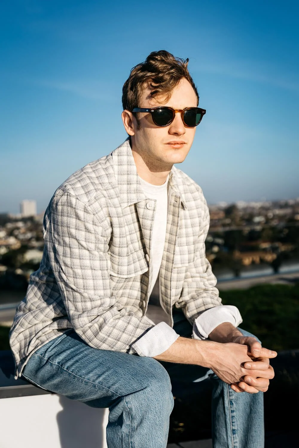 Man wearing jeans, white tee, and sunglasses sits on rooftop deck with Oakland skyline behind him.