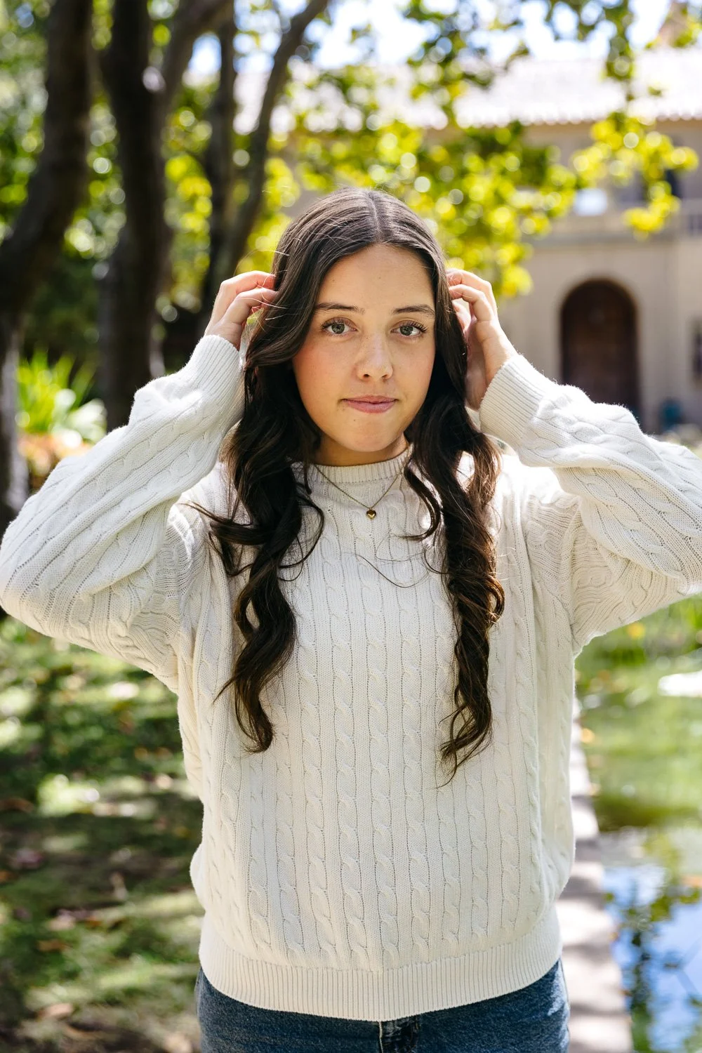 High school teen girl with long brown hair and white sweater stands under trees at Blake Gardens. Her hands are in her hair and she smiles at the camera with confidence. From outdoor senior photo shoot in Berkeley.