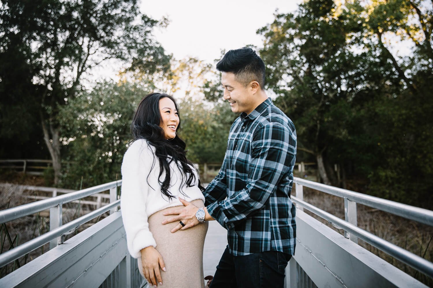 Husband places hand on pregnant belly of his laughing wife, shot during maternity session at Lafayette Reservoir.
