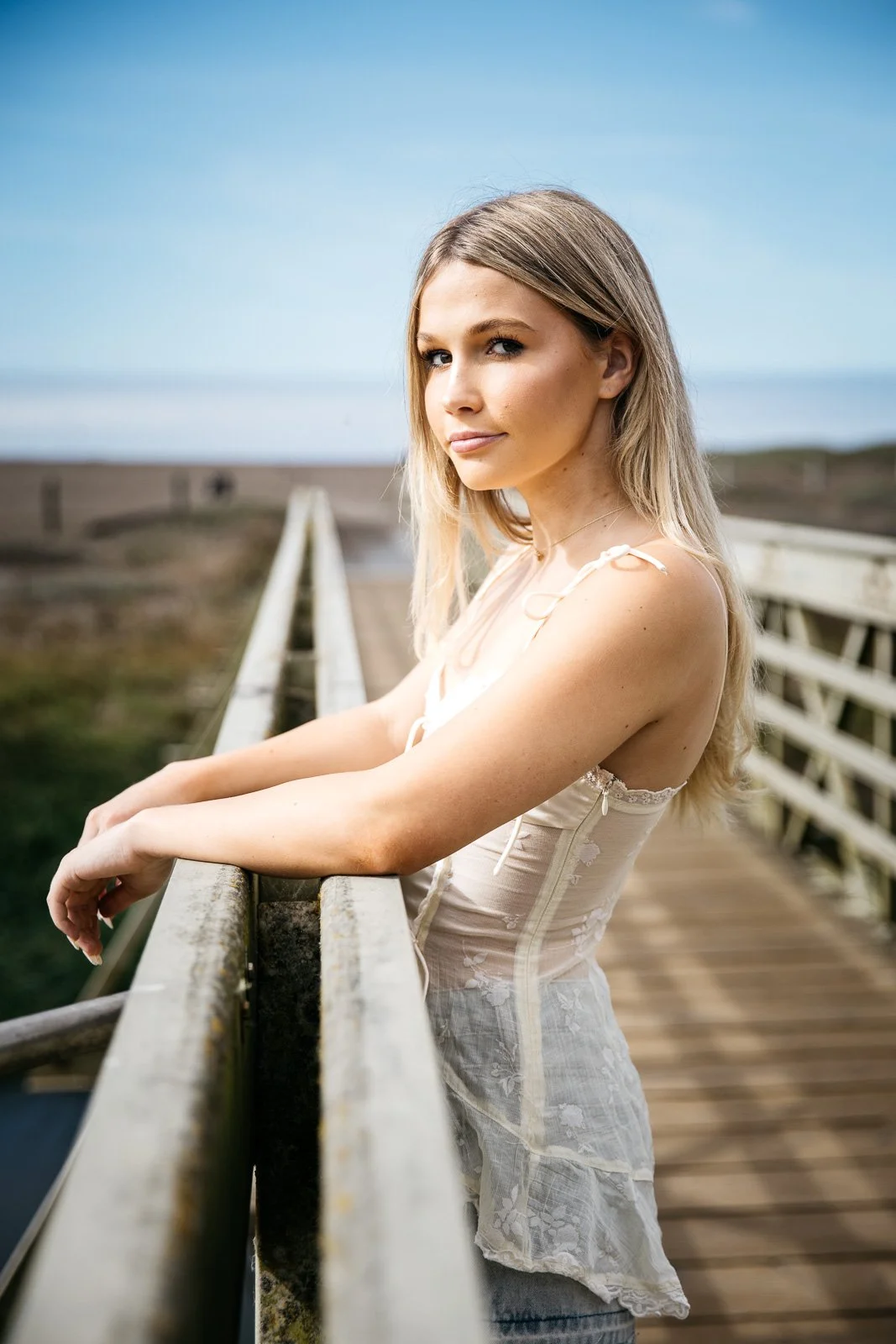 Teen girl with blond hair, white satin tank, and short denim skirt stands in profile with arms extended, on a footbridge at Rodeo Beach, shot during senior photo session.