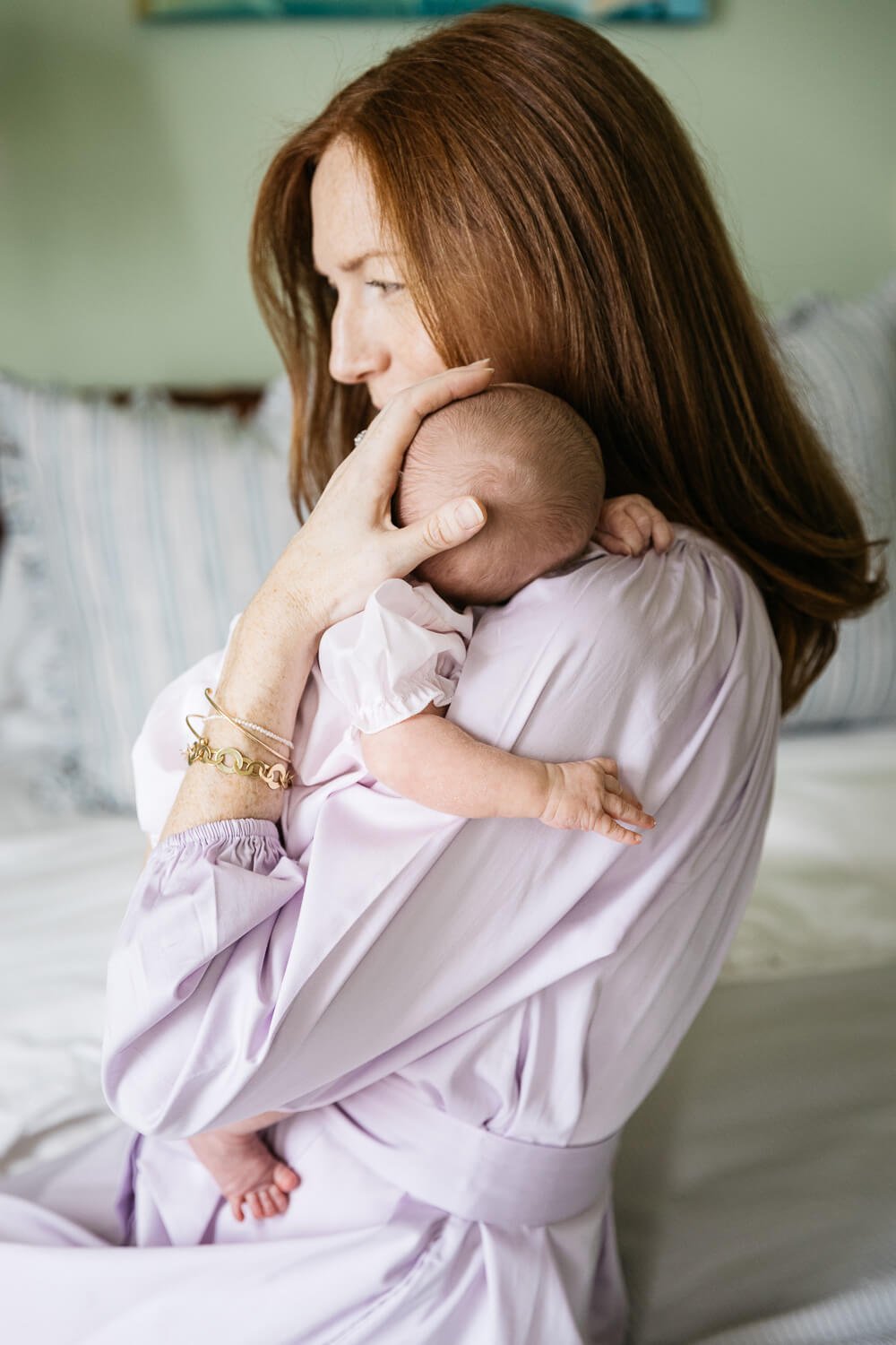 Mama in purple dress cradles newborn baby in room with pale green walls, shot at home in San Francisco.