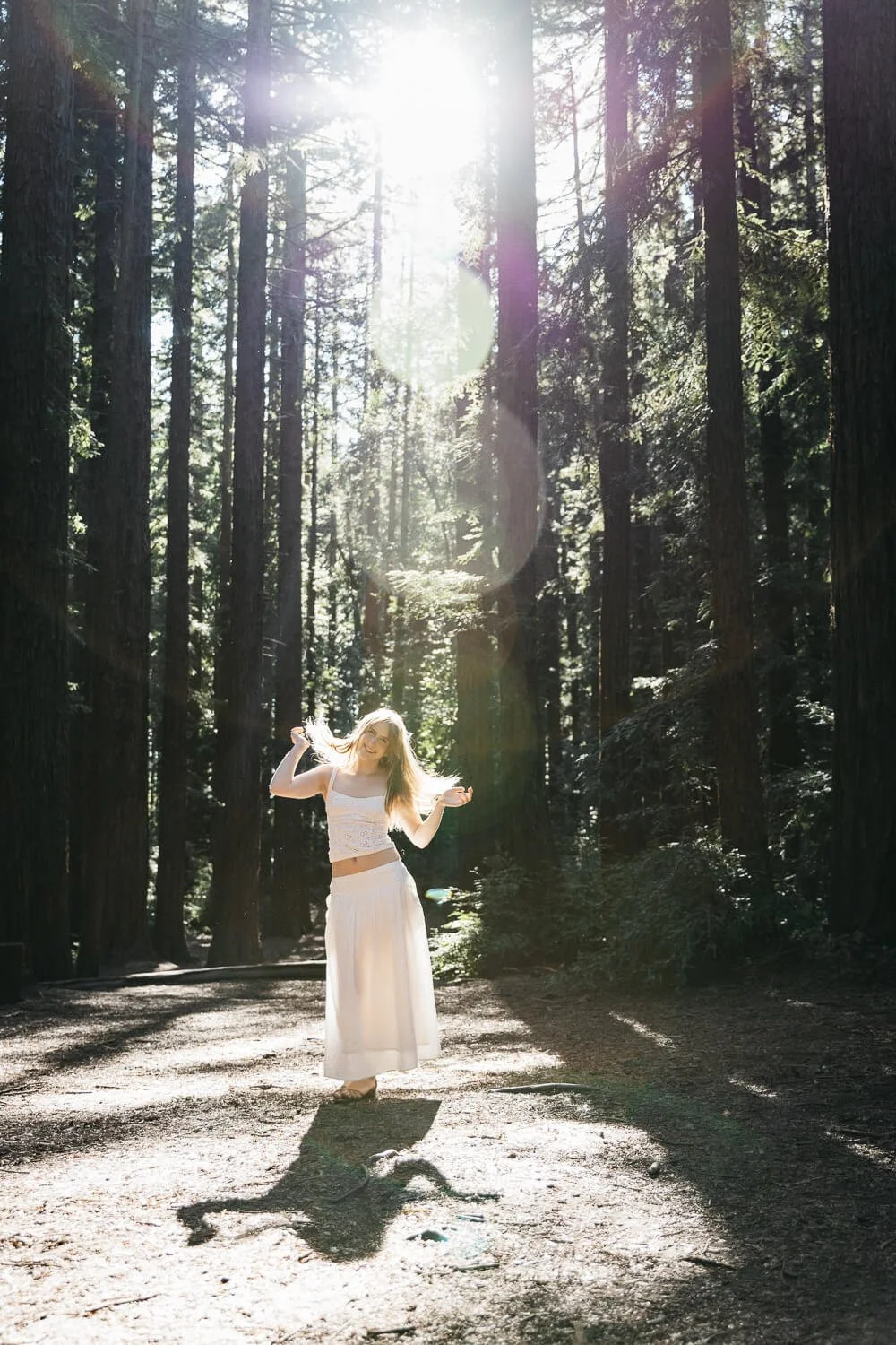 Teen in white skirt and tank top stands amongst redwoods and flips her hair as the light shines behind her. Shot during senior photo session at Redwood Regional in Oakland.