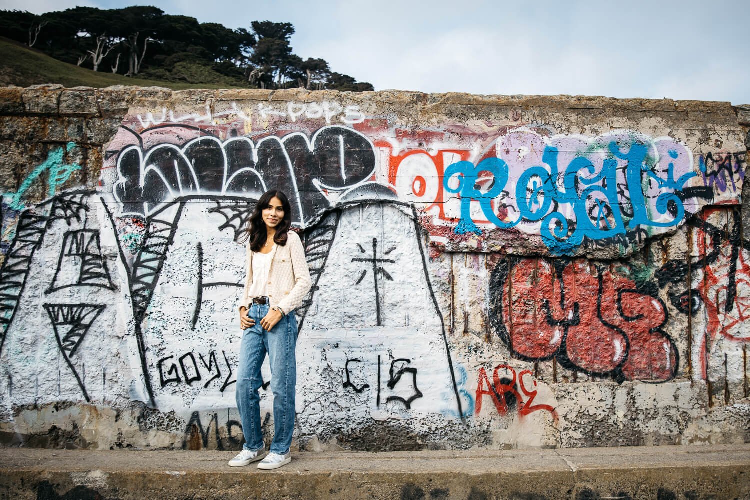 Teen in white sweater and jeans stands in front of grafitti wall at Sutro Baths in San Francisco.