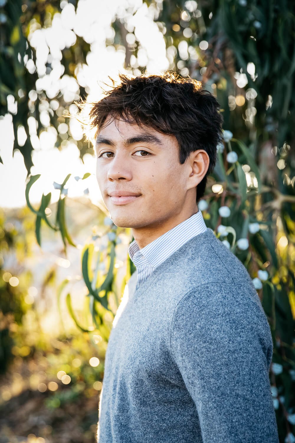 Teen boy stands smiling, backlit under a eucalyptus tree, shot during senior photoshoot at the Albany Bulb.