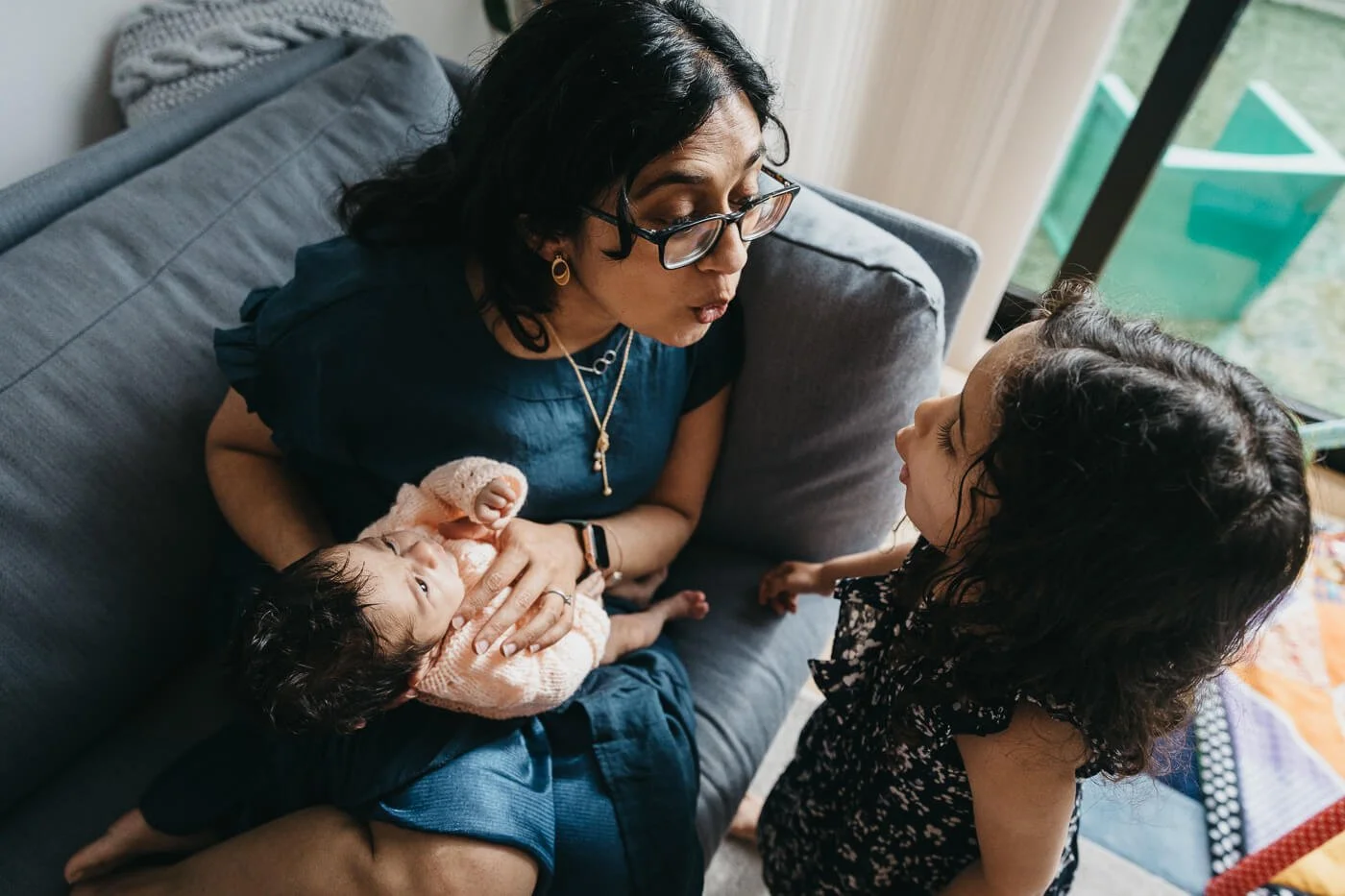 Mom holding newborn baby on living room couch, making silly faces with toddler daughter, shot during a lifestyle newborn photo session at home in San Francisco.