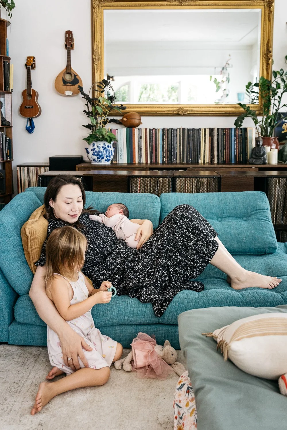 Mom reclines on turquoise colored couch, snuggling newborn baby and toddler daughter, who sits on the floor nearby.