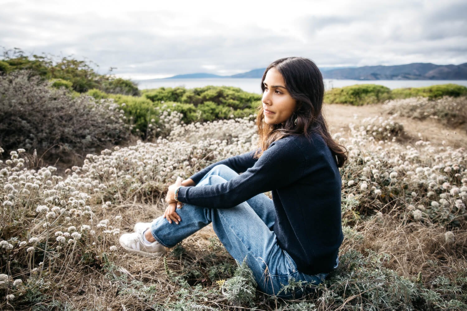 Teen with black hair and navy sweater gazes into sunset during senior photo session at Lands End.