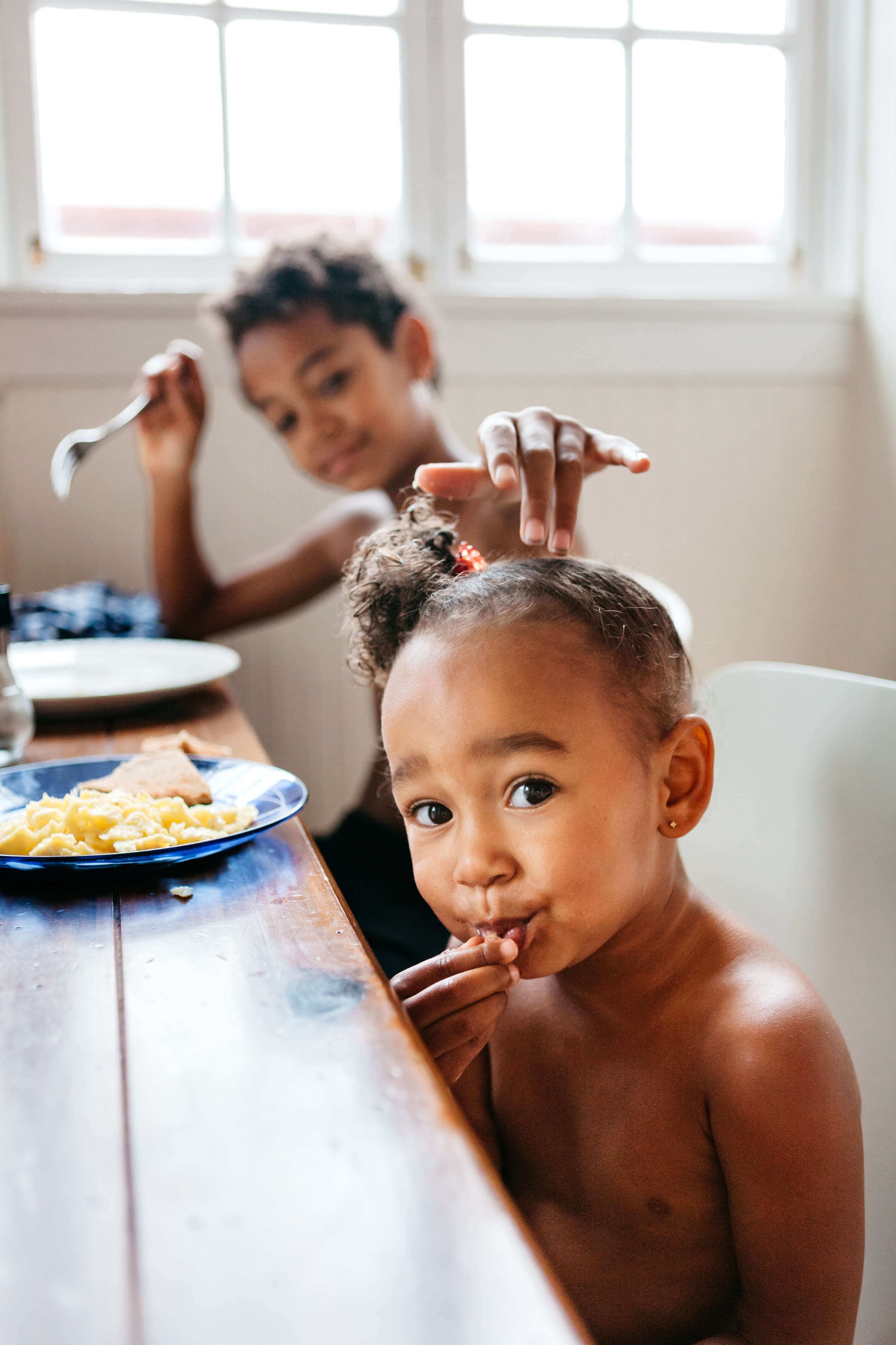 Toddler girl eats scrambled eggs off blue plate while sister pokes her from behind, shot during a home session.