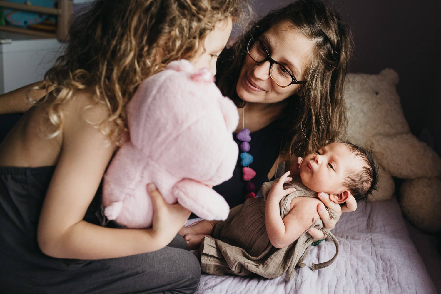 Mom holds newborn on bed with purple bedspread while older sister presents baby with pink CareBear.