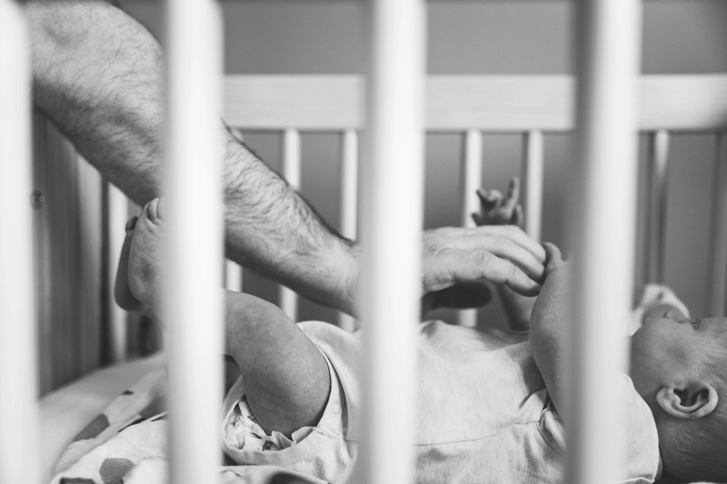 Black and white image of dad's arm reaching into crib to calm newborn baby, shot during a newborn session in Oakland.