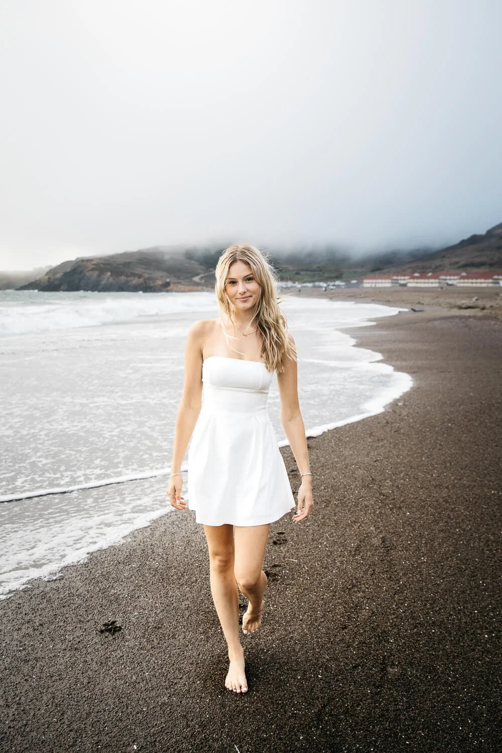 Barefoot teen girl with long blond hair and short white dress walks in surf at foggy Rodeo Beach, shot during senior photo session.