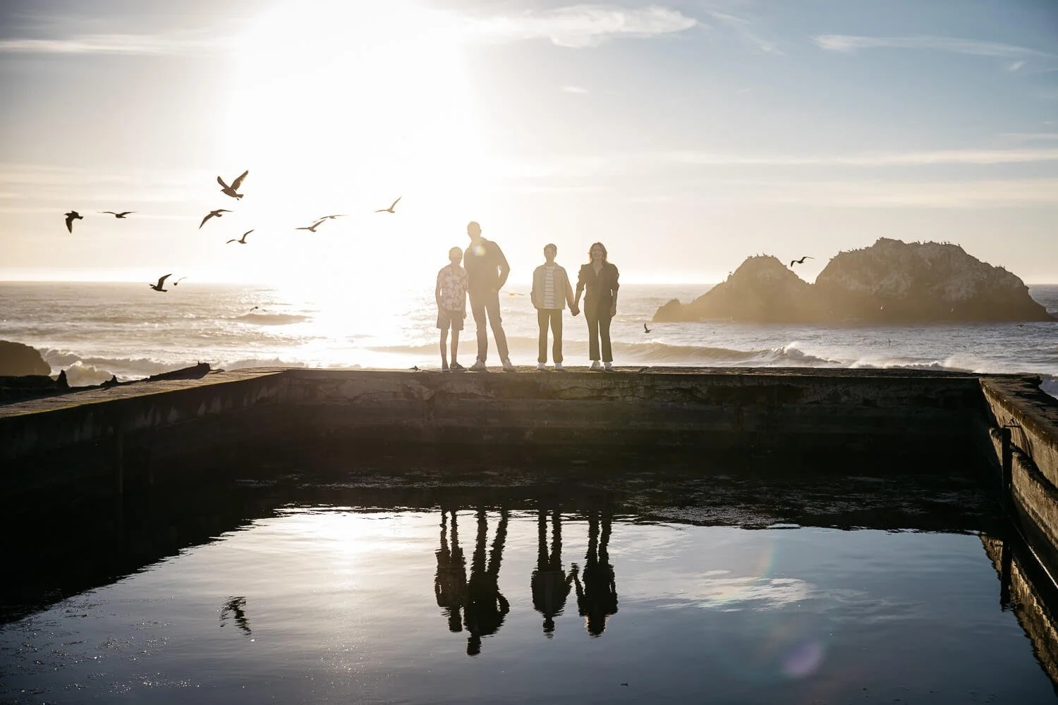A family of 4 stands on a ledge at the Sutro baths in San Francisco. The sun sets into the ocean behind them and gives a golden reflection in the water. Pelicans fly overhead. Taken during an outdoor mini family photo shoot.