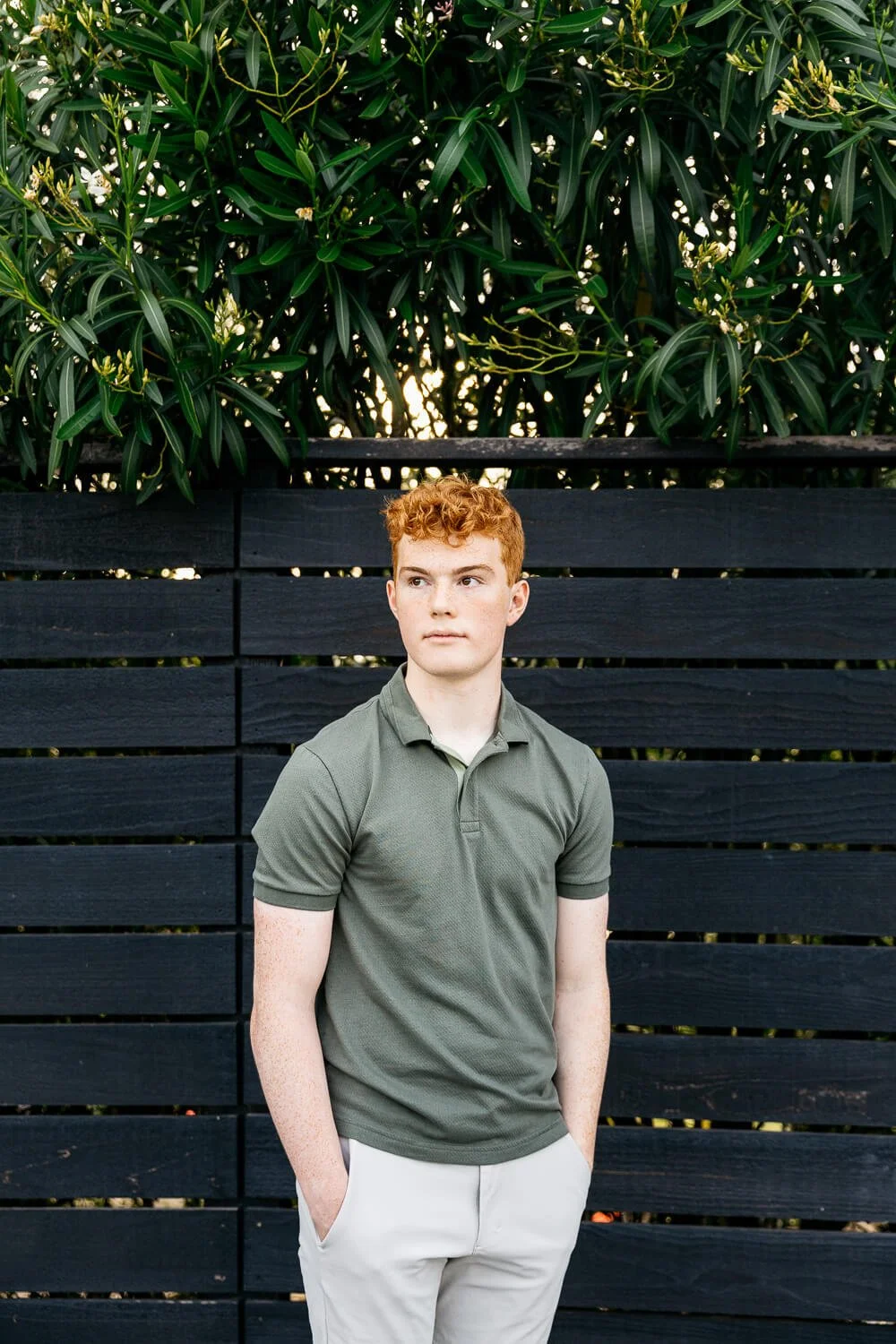 Senior portrait of redheaded young man in green polo against a black fence with oleander bushes, shot in Orinda, CA.