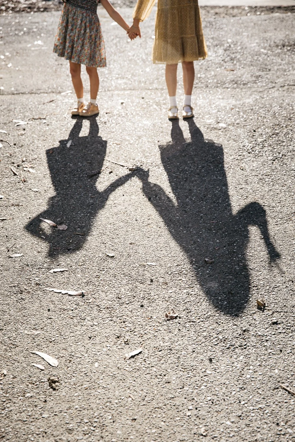 Two sisters stand holding hands in bright sunlight as their shadows fall on the pavement.