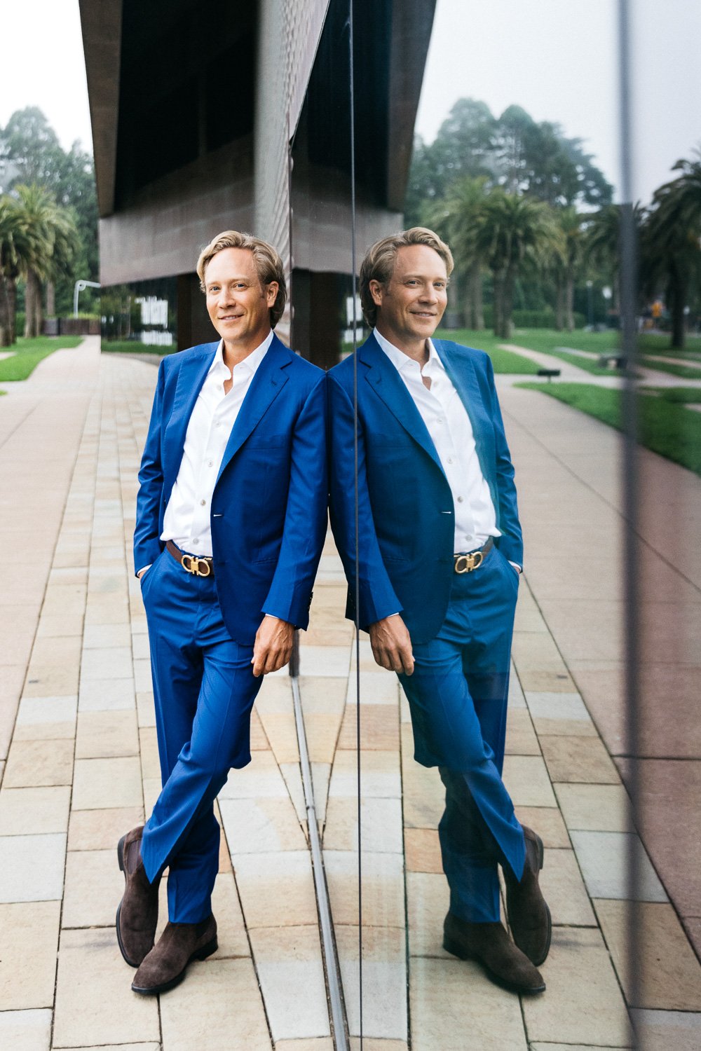 Blonde man in blue suit, posed against wall of the deYoung Museum in San Francisco. He stands with crossed legs, one hand in pocket, shoulder leaning against the building, smiling. He looks at ease and confident.