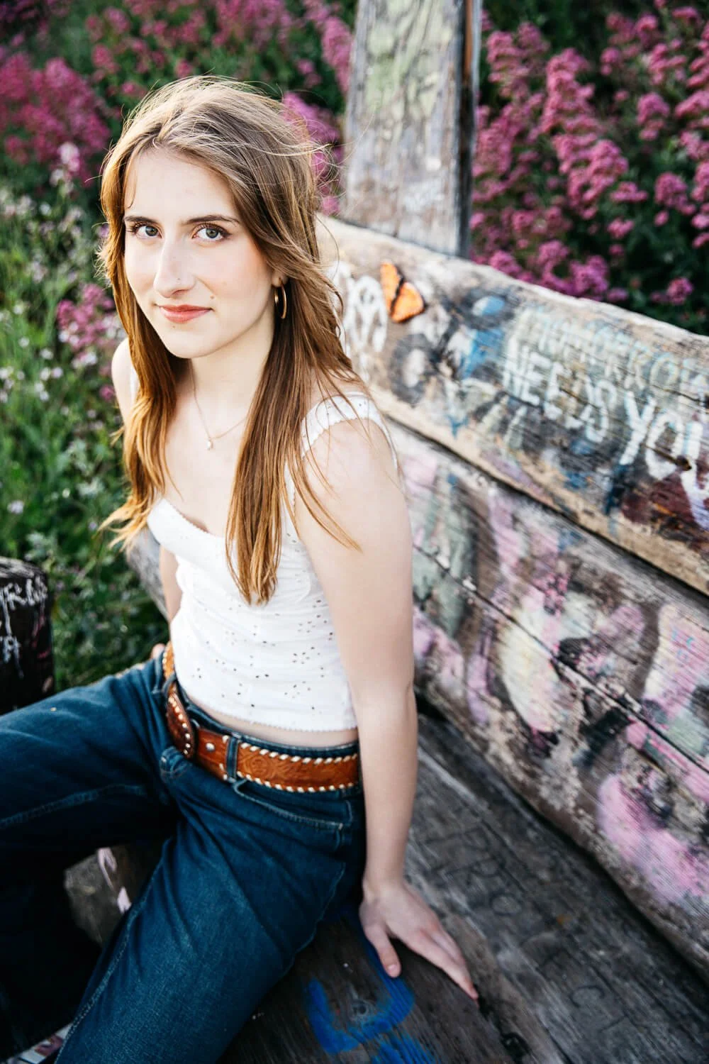 Berkeley senior photographer captures high school senior leaning against a graffiti-covered wooden structure at the Albany Bulb, surrounded by pink wildflowers, wearing a white eyelet top and western belt.