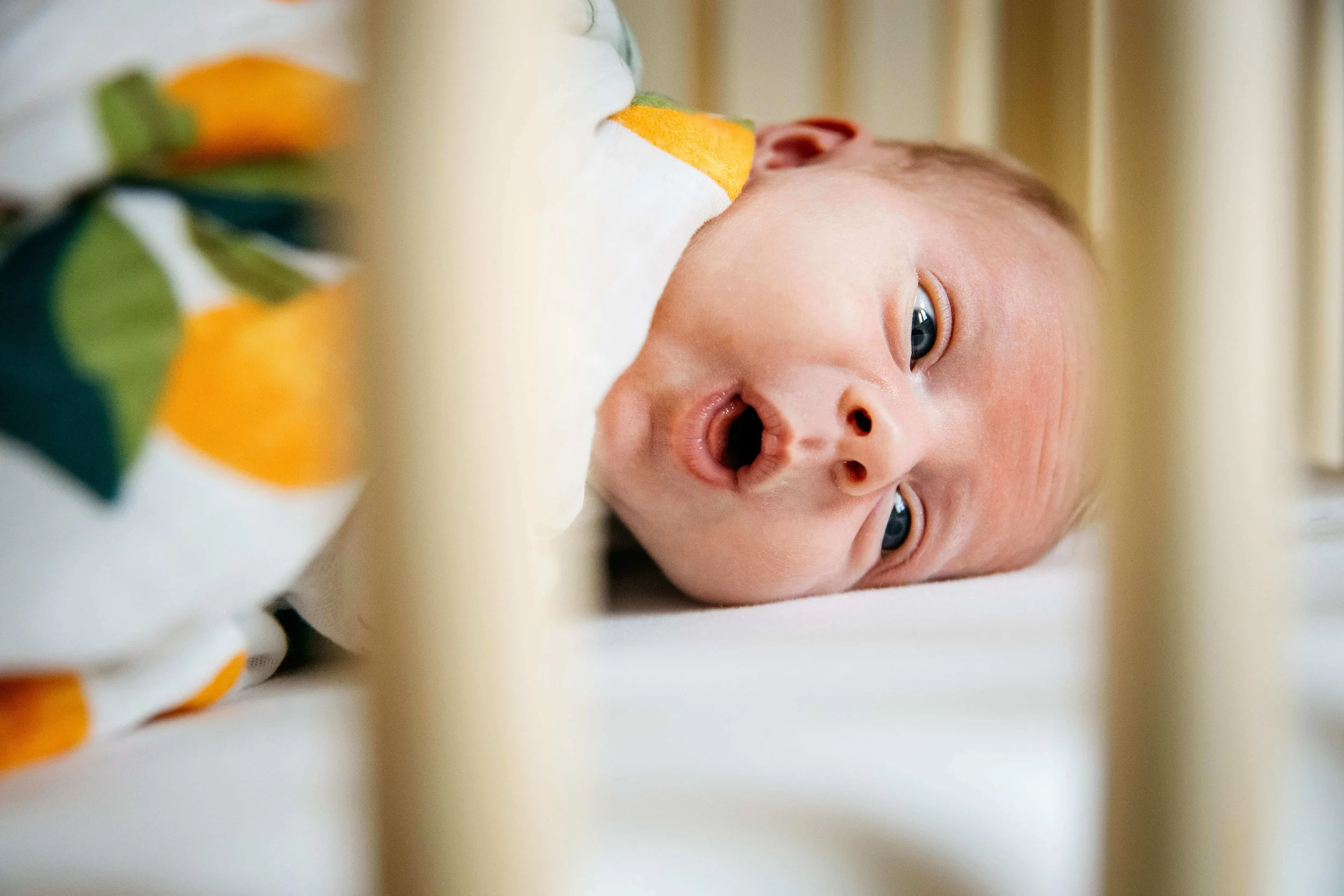 Close up, through the bars of a crib, of a newborn baby wrapped in a blanket with bright oranges, shot at home in Berkeley.