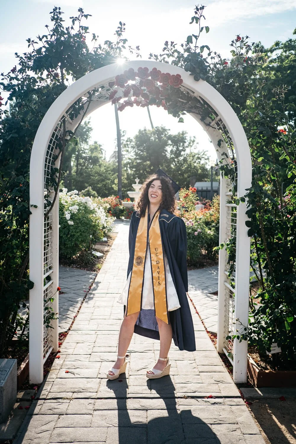 Graduating girl wears UC Davis cap and gown and stands under an archway covered in green vines and roses.