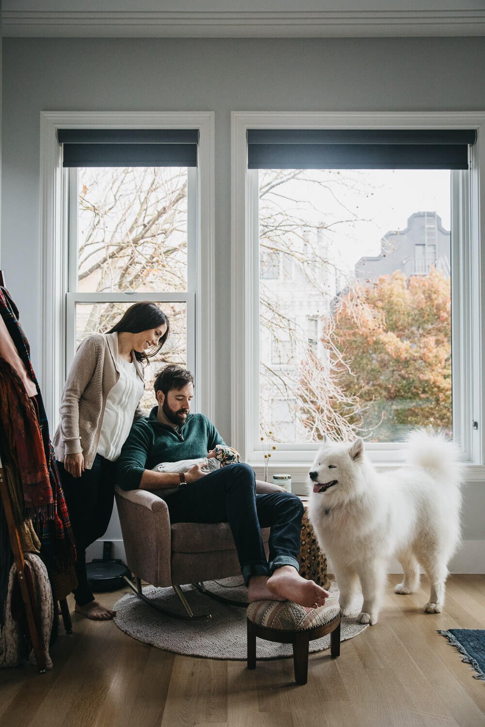 Mom and Dad gaze down at newborn while sitting near large window with large white, fluffy dog standing nearby.