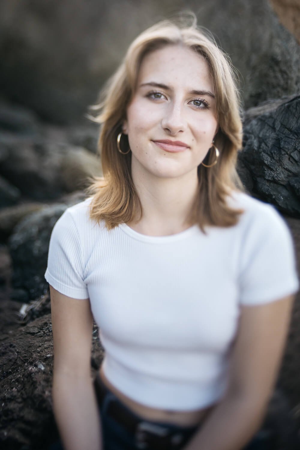 High school senior in white t-shirt smiles while sitting on beach rocks at Rodeo Beach in Marin Headlands.