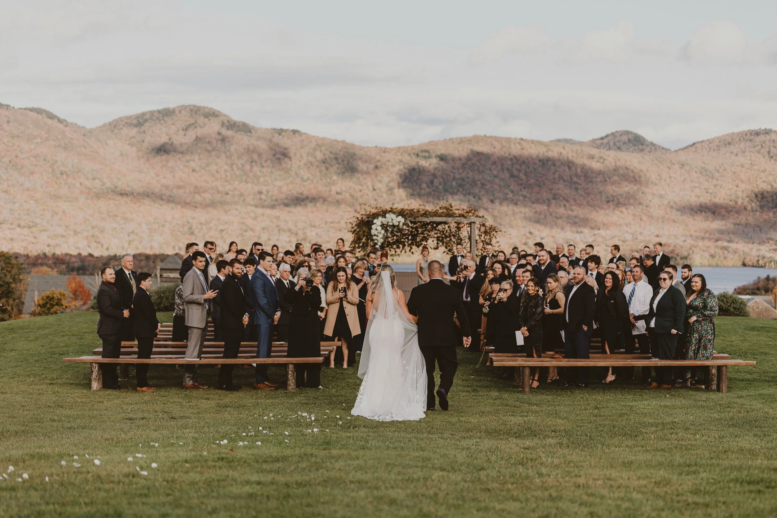 Bride and groom walking down the aisle during an outdoor wedding ceremony with guests in attendance and scenic mountains in the background.